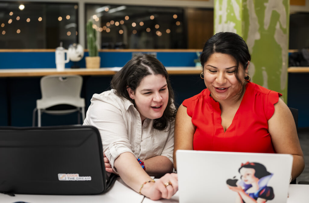 Two people sit in front of laptops, one assisting the other.